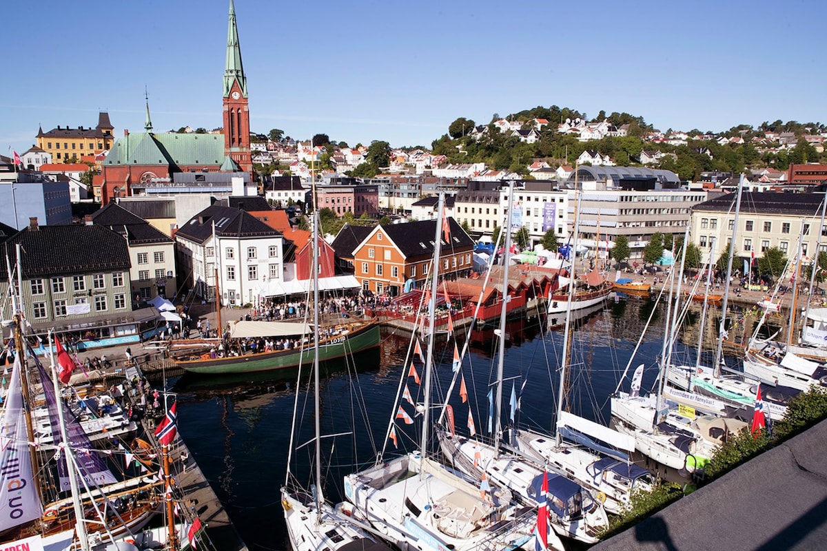 A vibrant harbor scene features a mix of colorful boats and yachts docked along the waterfront. A bustling promenade is visible, with people enjoying the lively atmosphere. The backdrop includes charming buildings and a steeple of a church, set against a clear blue sky.
