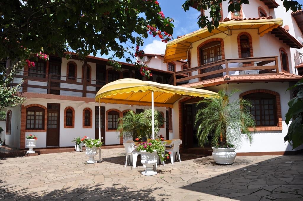 A courtyard features a yellow canopy providing shade over a seating area with white chairs. Surrounding flower pots add color to the stone-paved space. The two-story building showcases traditional architecture with wooden accents and balcony areas, under a clear blue sky.