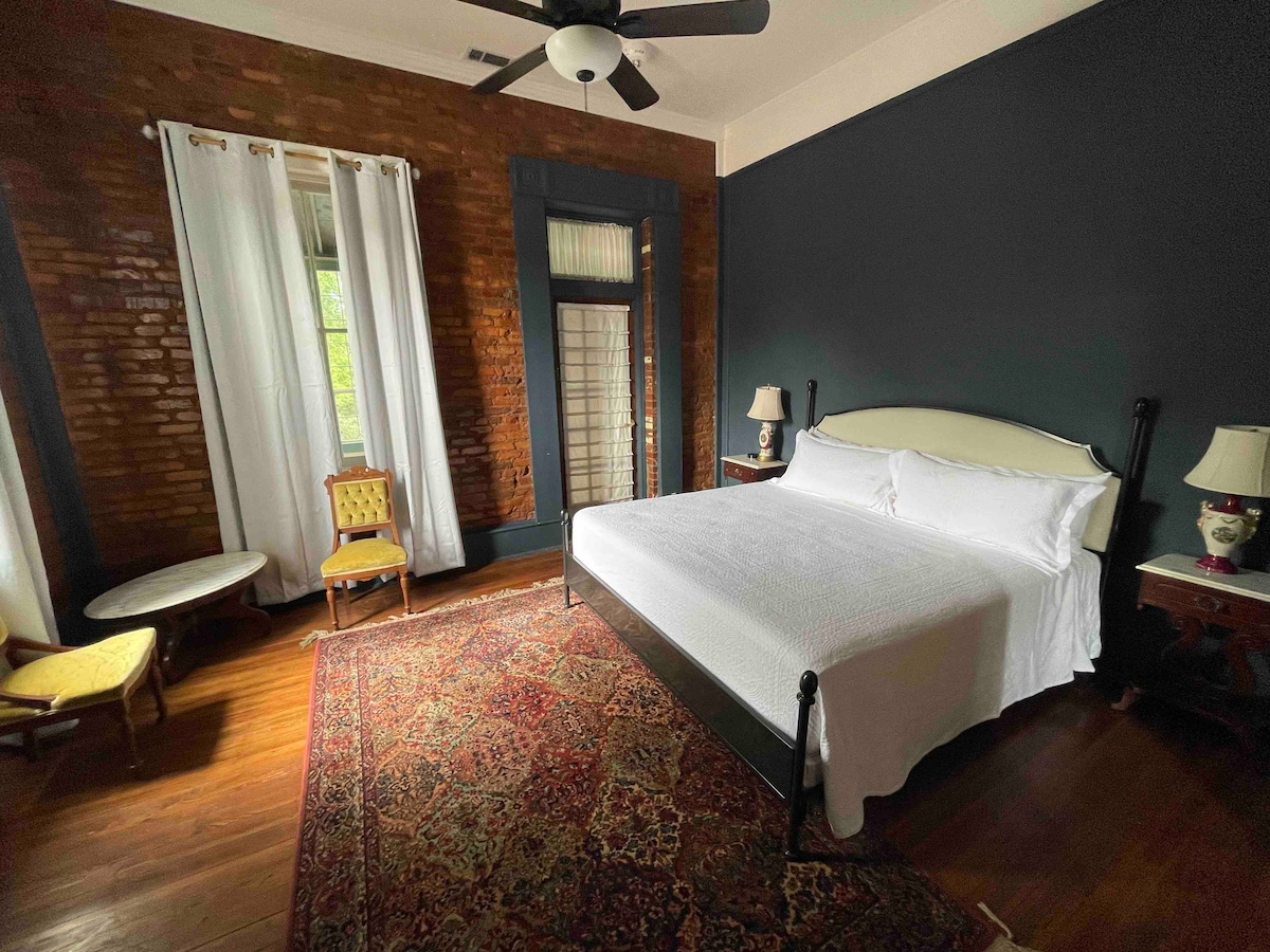 A spacious bedroom features a large bed dressed in crisp white linens. Exposed brick walls add character, while large windows are adorned with light curtains. A vintage chair and a small round table are positioned in the corner, complemented by bedside tables with lamps.