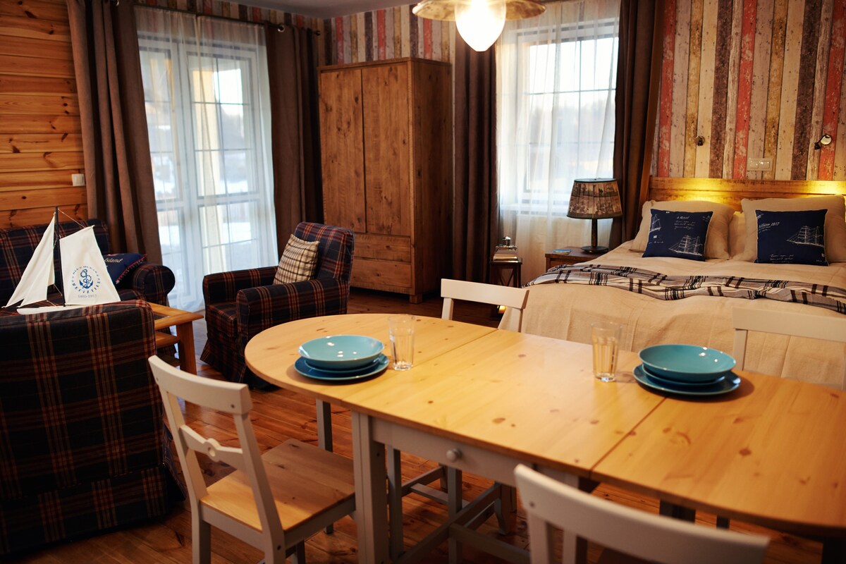 A cozy room is presented with wood-paneled walls and patterned wallpaper. A spacious bed is paired with a plaid armchair and a wooden dining table set for four. Natural light fills the space through large windows dressed in curtains, enhancing the warm atmosphere.