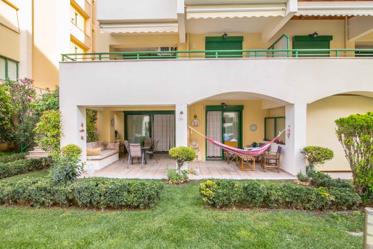 An outdoor sitting area features a hammock suspended between two pillars, surrounded by well-kept greenery. Adjacent dining furniture is visible alongside a comfortable lounge area, with the facade of the home displaying large glass doors that invite natural light into the space.