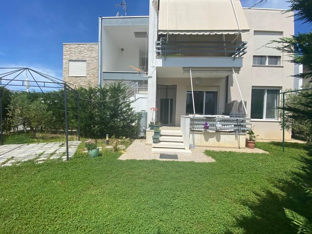 A garden area features a well-maintained lawn surrounded by greenery. The building showcases a spacious terrace with railing, offering access through sliding glass doors. A covered seating area is visible, providing shade and comfort, while stone pathways lead to the entrance.