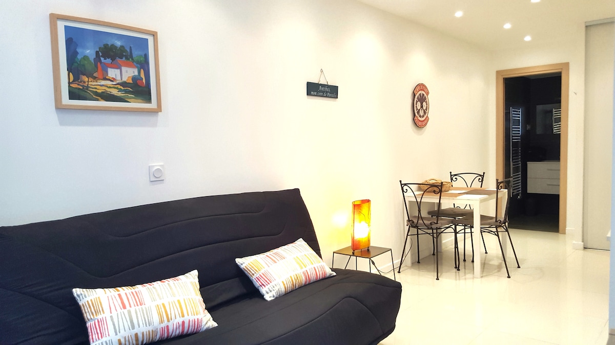 A modern studio features a sleek black sofa with decorative pillows, illuminated by a warm floor lamp. A wooden dining table with four chairs is positioned in the background, complementing the bright, neutral walls and contemporary ambiance.