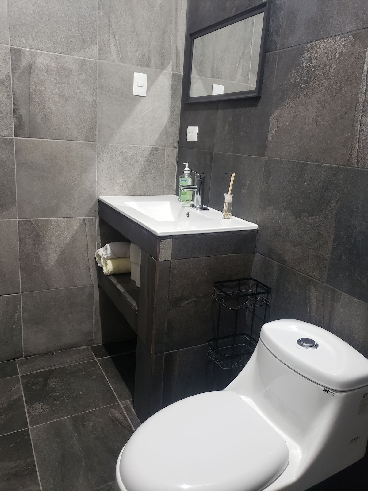 A modern bathroom features dark gray tile walls and flooring. A sleek vanity with a rectangular sink is positioned against the wall, complemented by a mirror above. Toiletries are neatly arranged on the countertop, while a toilet and a black wire basket are also present.