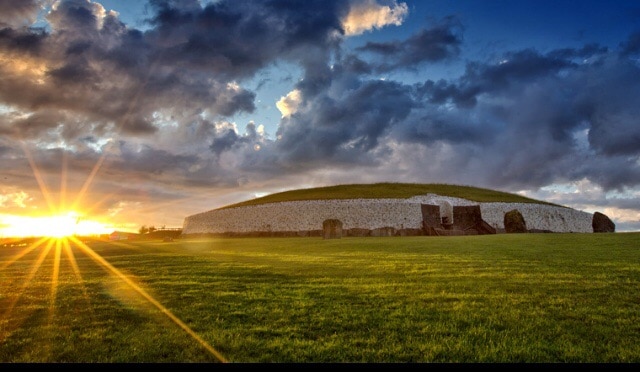 An ancient passage tomb, known for its distinct circular stone structure, is illuminated by the soft glow of the rising sun. The surrounding grassy landscape enhances the monument's historic presence against a dramatic sky filled with clouds.