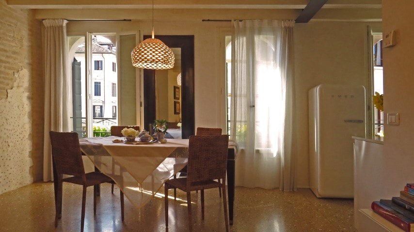 A dining area features a light wooden table set for two, accompanied by four woven chairs. Large windows with soft curtains allow natural light to fill the room, providing views of the cityscape outside. A white refrigerator is visible in the corner.