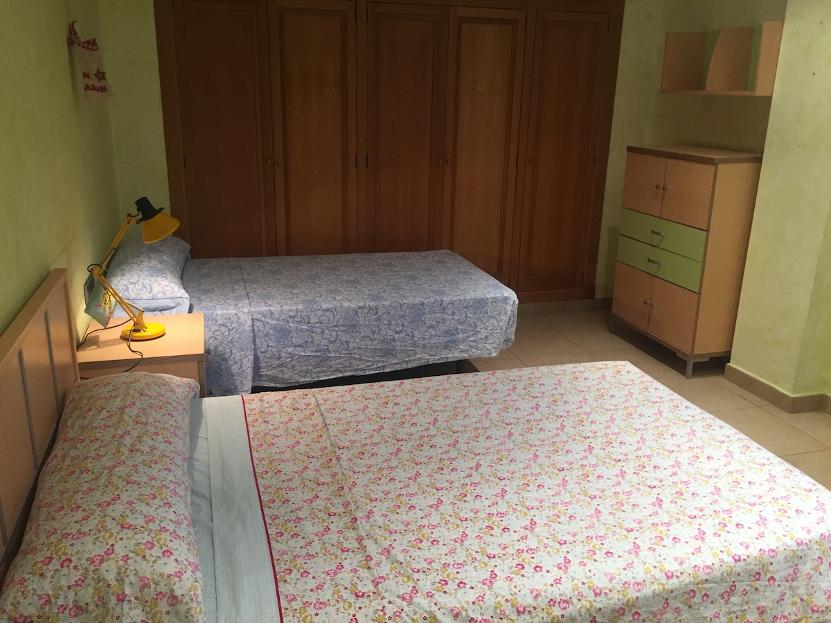 A room is equipped with two beds, one larger and one smaller, covered in floral and patterned bedding. A light-green lamp provides illumination on a bedside table. Ample storage is available in wooden wardrobes and a matching dresser with colorful drawers.