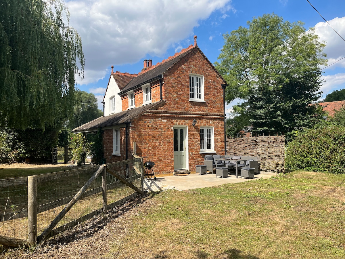 The cottage, with its charming red brick exterior, is framed by greenery. A spacious patio area features outdoor seating arrangements, alongside a barbeque. The surrounding garden is grassy and offers a serene setting, with a fence marking the property boundary.