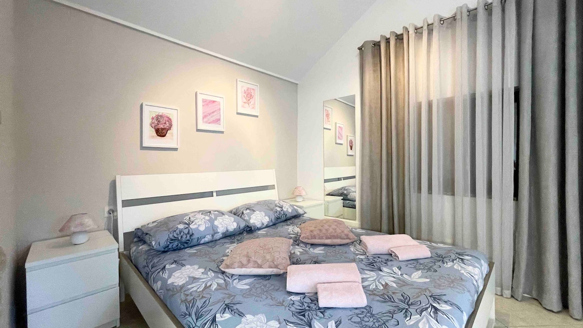 A bright bedroom features a spacious bed adorned with floral-patterned bedding. Soft pink towels and decorative cushions are arranged neatly on the bed. Light-colored walls are complemented by pale curtains, and framed artwork adds a touch of color and detail to the inviting space.