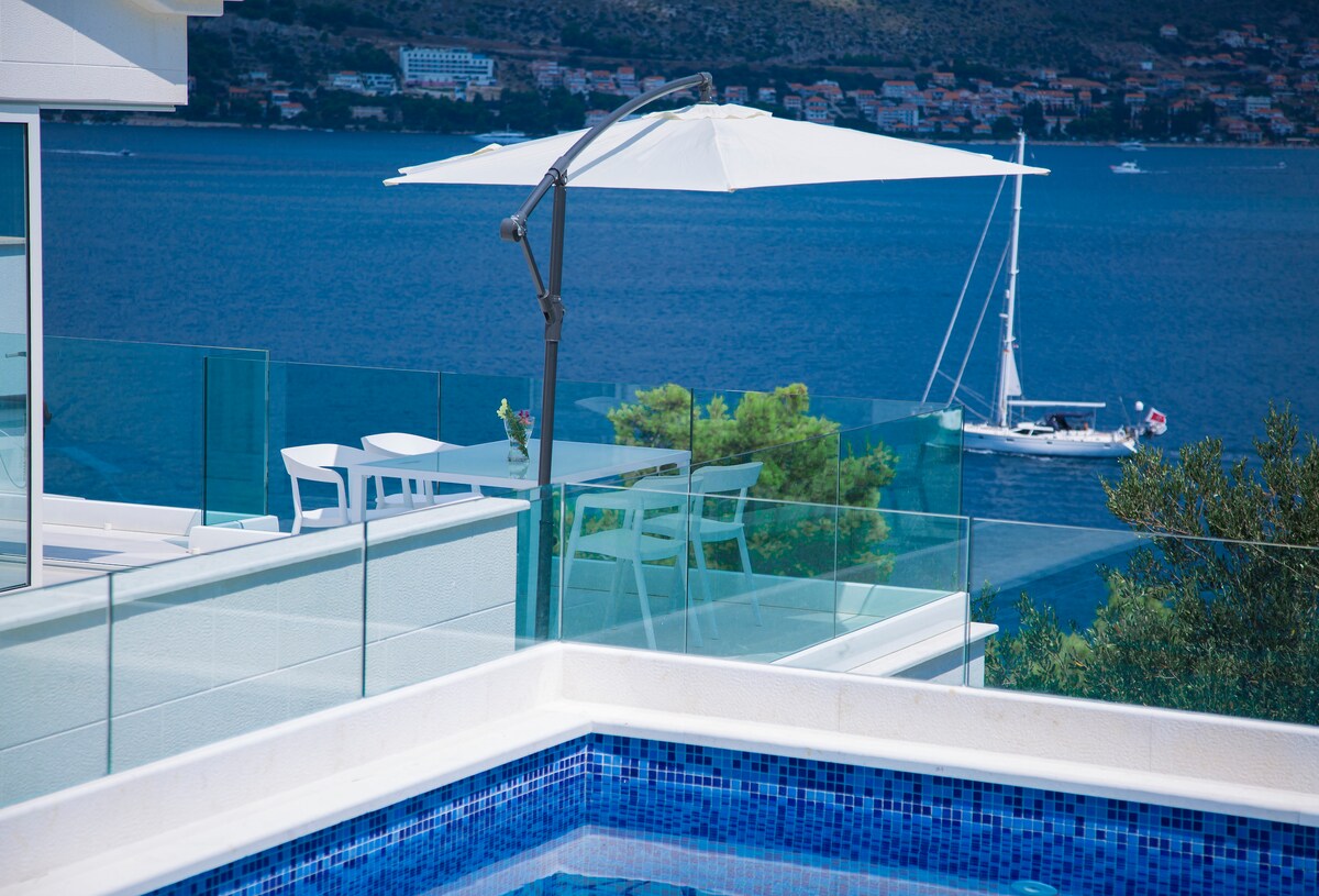 A terrace with a sleek white umbrella provides shade over a glass table and chairs. The view features a sailboat on the calm sea and distant coastal scenery, framed by the clear blue sky and verdant foliage.