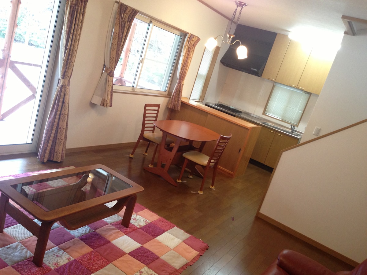 An open living space features a round dining table with two chairs, complemented by a cozy sofa and a glass coffee table. Natural light pours in through large windows, illuminating the wooden flooring and simple kitchen area in the background.