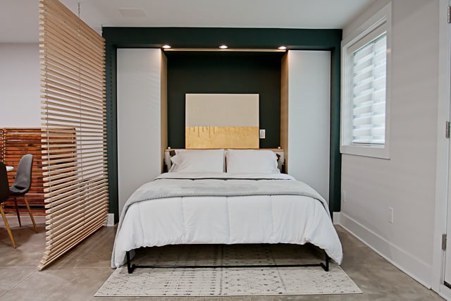 A spacious studio is depicted, featuring a comfortable bed positioned against a dark accent wall. Decorative lighting highlights a modern artwork above the bed. A textured rug lies beneath, while a wooden partition offers a sense of separation from the dining area in the background.
