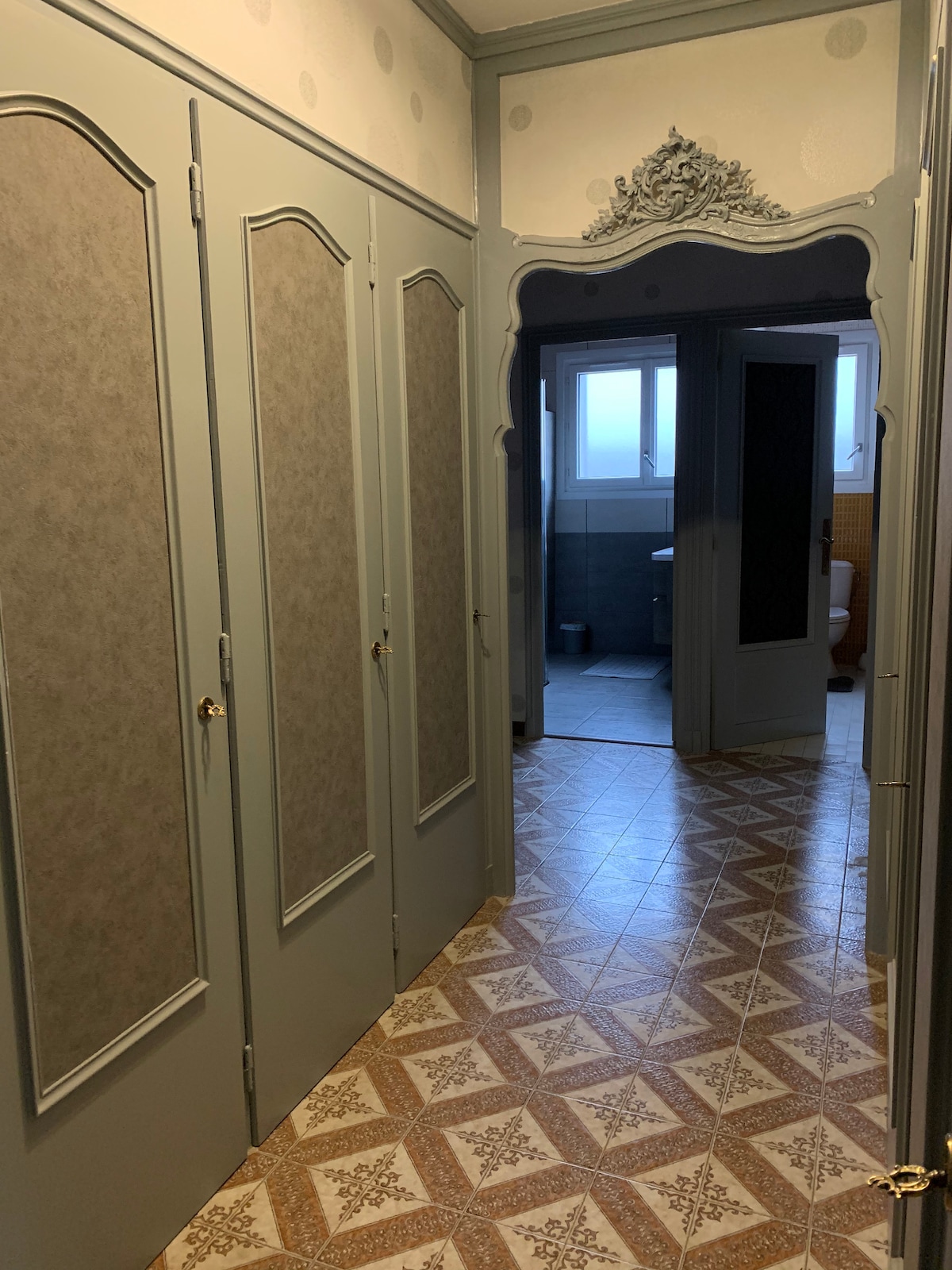 A hallway is shown with ornate grey cabinetry along one side. The tiled floor features a patterned design, while a doorway at the end leads to a bathroom area. Soft light enters through a window, highlighting the decorative molding on the walls.