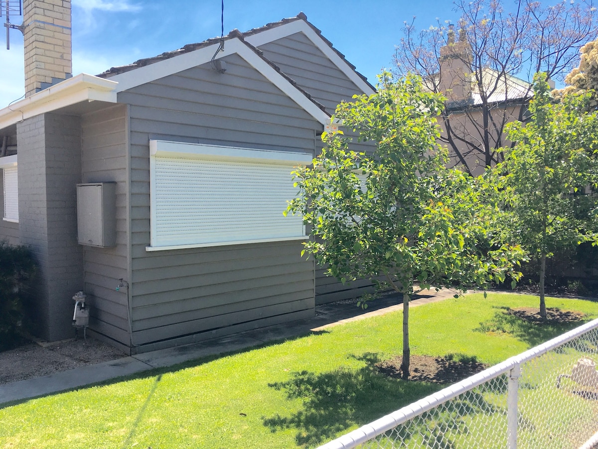 The exterior of a modern, single-story building is shown, featuring light gray cladding with white trim. The well-maintained lawn is punctuated by two small trees. Stylish roller shutters cover the windows, providing privacy and shade. A fence surrounds the property, ensuring security.