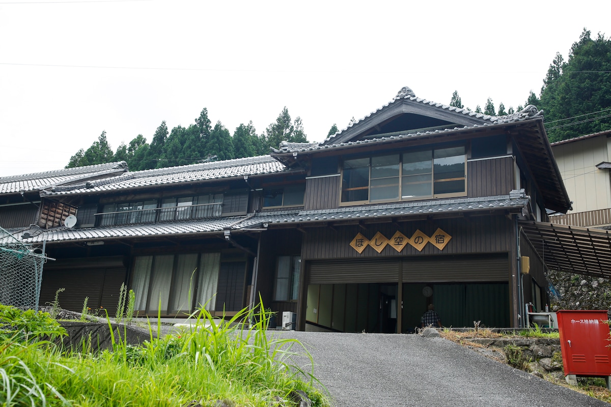 A traditional two-story building features dark wooden siding and a tiled roof. Large windows provide a view of the surroundings, while a serene pathway leads up to the entrance. Lush greenery and trees frame the structure, contributing to the natural landscape.