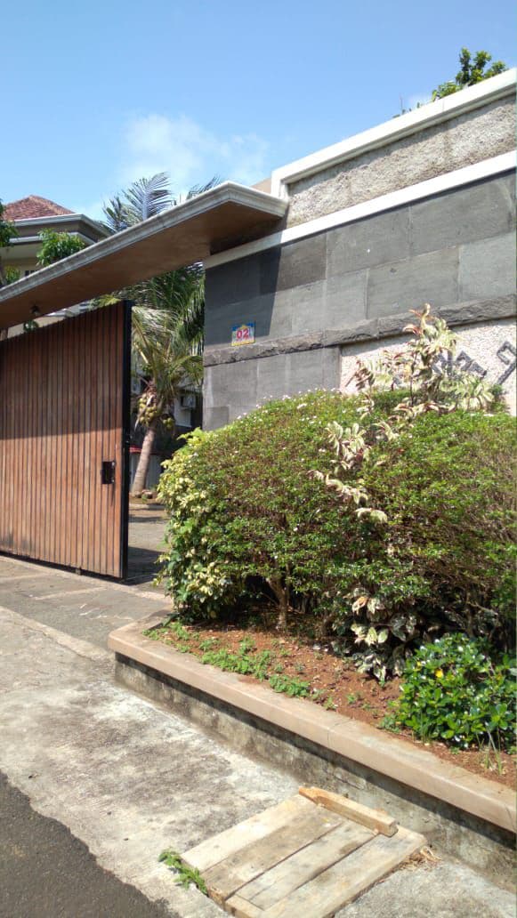 A wooden gate stands partially open, revealing a pathway lined with lush greenery. A stone wall provides a textured backdrop, while a vibrant garden with shrubs and plants offers a welcoming approach to the property. The clear sky suggests a sunny day.