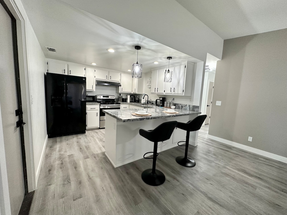 A modern kitchen features white cabinetry and a spacious granite countertop with two black bar stools. Stainless steel appliances, including a fridge and oven, are visible. Natural light brightens the area, enhancing the contemporary feel of the space.