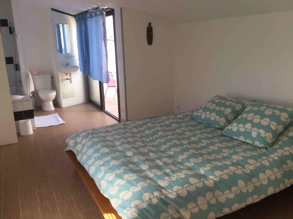 A bright sleeping area features a spacious bed adorned with a blue and white patterned duvet and two matching pillows. Light streams through large glass doors that open to a small terrace. A private bathroom is visible, equipped with modern fixtures.