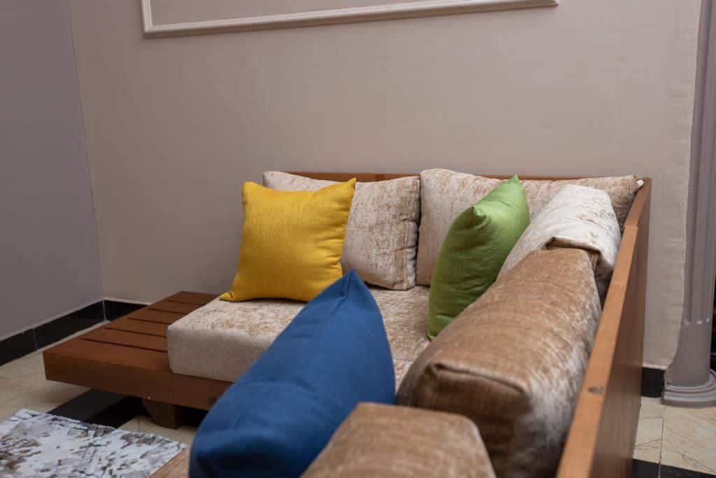 A modern, wooden seating area is displayed, featuring a spacious, low-profile couch adorned with decorative cushions in vibrant colors: yellow, green, and blue. The neutral fabric of the couch complements the surrounding walls and flooring, creating a comfortable and inviting ambiance.