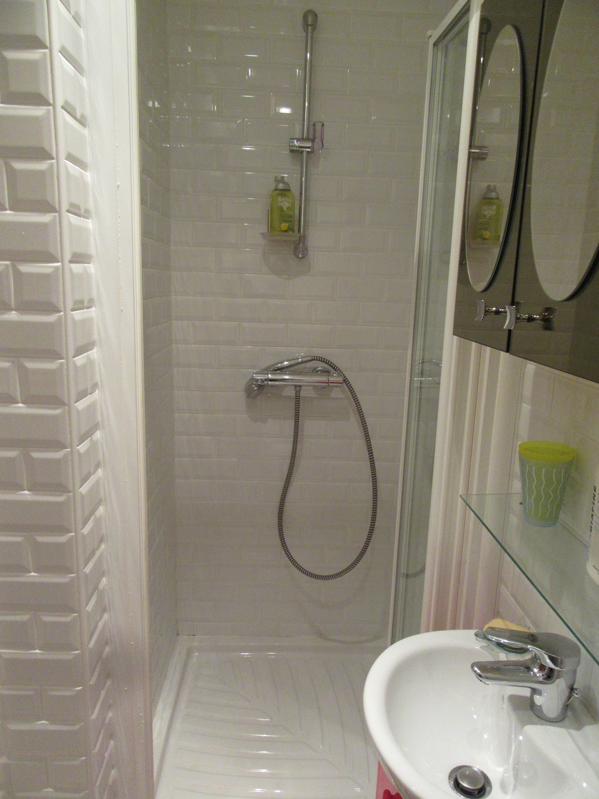 A compact bathroom area is presented, featuring a tiled shower stall with a modern faucet and adjustable showerhead. A glass shelf holds toiletries above a small sink, while a decorative mirror is mounted on the wall, reflecting a clean and bright atmosphere.