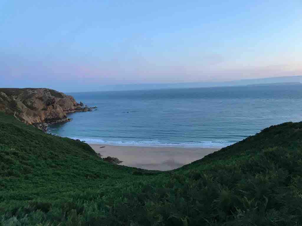 A serene coastal view is captured, showcasing gentle waves lapping against a sandy beach framed by lush green hills. The sky transitions from soft pinks to light blues, indicating twilight. The image suggests a tranquil atmosphere near this picturesque shoreline.
