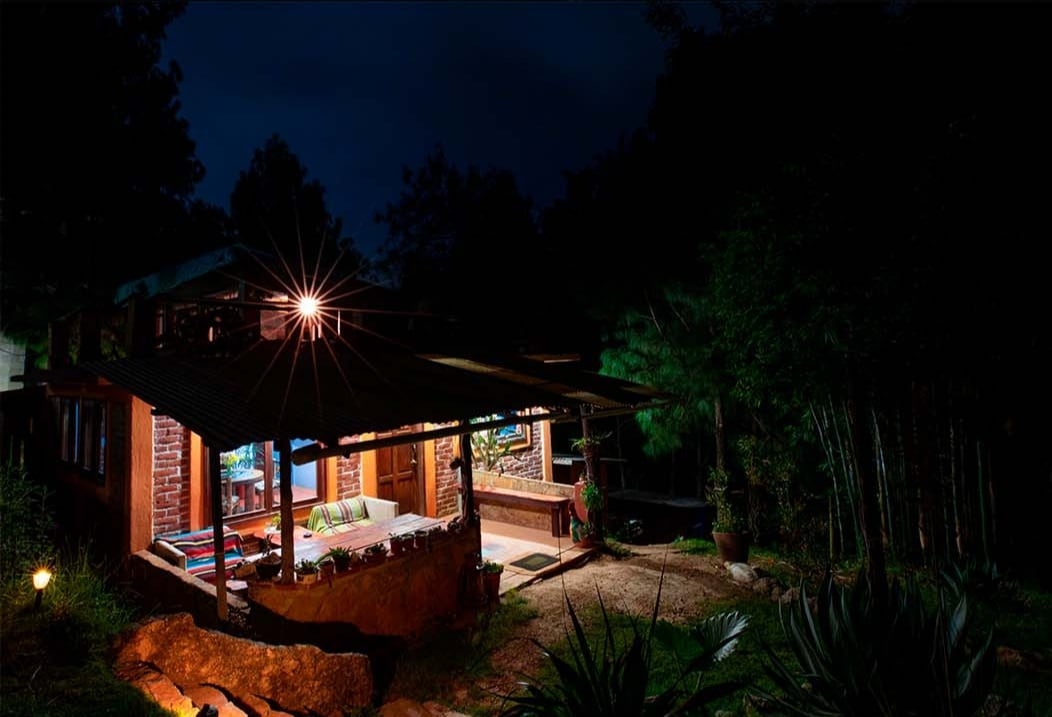 The exterior of a rustic cabin is illuminated at night, showcasing a welcoming porch with comfortable seating. Surrounding greenery adds to the natural ambiance, while soft lighting highlights the brick and wood features, creating an inviting atmosphere.