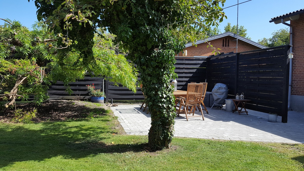A treed garden area features a shaded seating space, equipped with a wooden dining table and folding chairs. A grill is positioned nearby, and a paved patio area is visible. Lush greenery surrounds the space, providing a sense of privacy and tranquility.