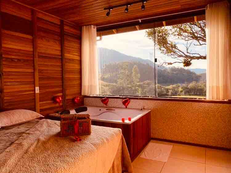A spacious bedroom features wooden walls and large windows framing views of distant mountains. A small jacuzzi is situated near the window, and soft lighting enhances the serene atmosphere. Light drapes complete the room, allowing natural light to filter in.