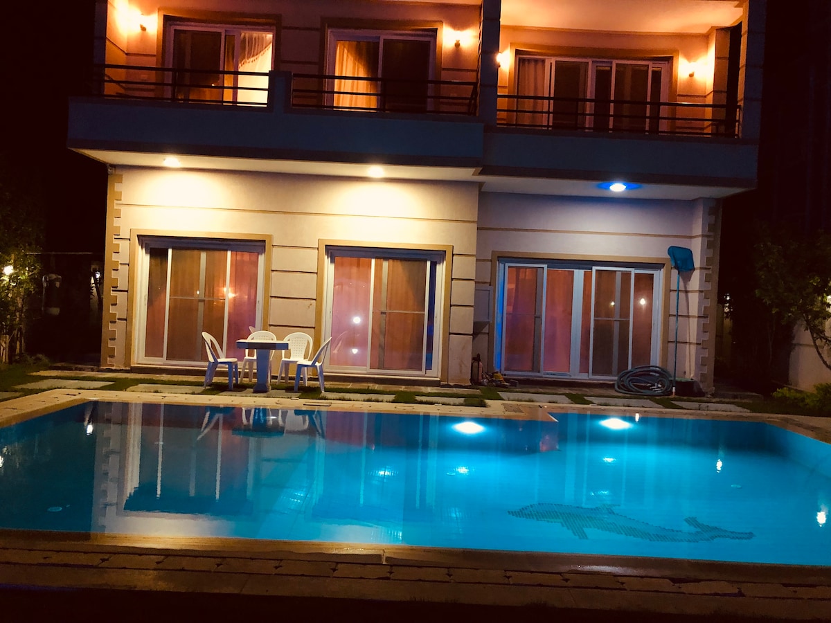 A two-story villa is illuminated at night, showcasing large glass doors that open to a sparkling swimming pool. Outdoor seating for four is positioned beside the pool, reflecting soft lighting from the villa. The tranquil water is calm, providing a serene ambiance.