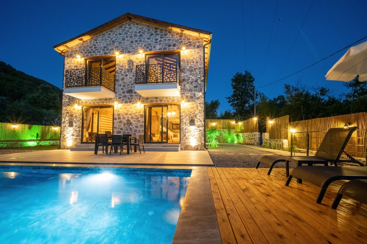The villa’s stone exterior is illuminated at night, showcasing a spacious pool area and outdoor seating. Large glass doors provide access to the interior, with inviting lighting highlighting the natural textures. A well-maintained garden can be seen in the background.