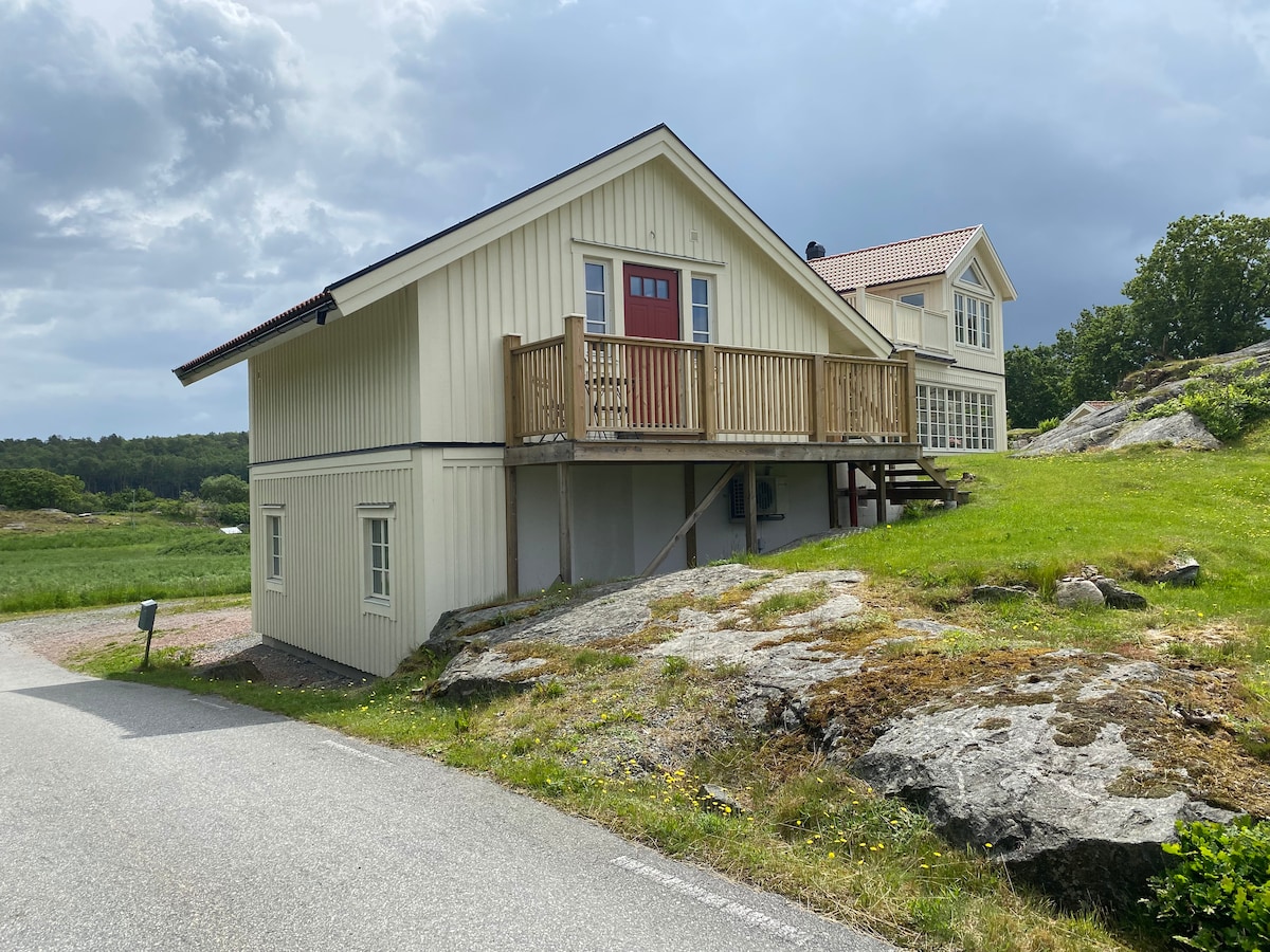 A charming two-story house is situated on raised ground, with a wooden deck visible at the entrance. A grassy area surrounds the property, while rocky terrain is present nearby. The exterior features light-colored siding and large windows, contributing to a spacious appearance.