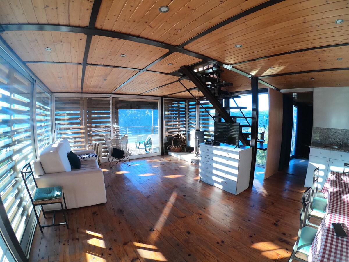 A spacious interior space features wooden ceilings and flooring, accented by large windows allowing natural light. Comfortable seating options include a white sofa and chairs. A staircase leads to the upper level, while a dining area with a checkered tablecloth is visible in the background.