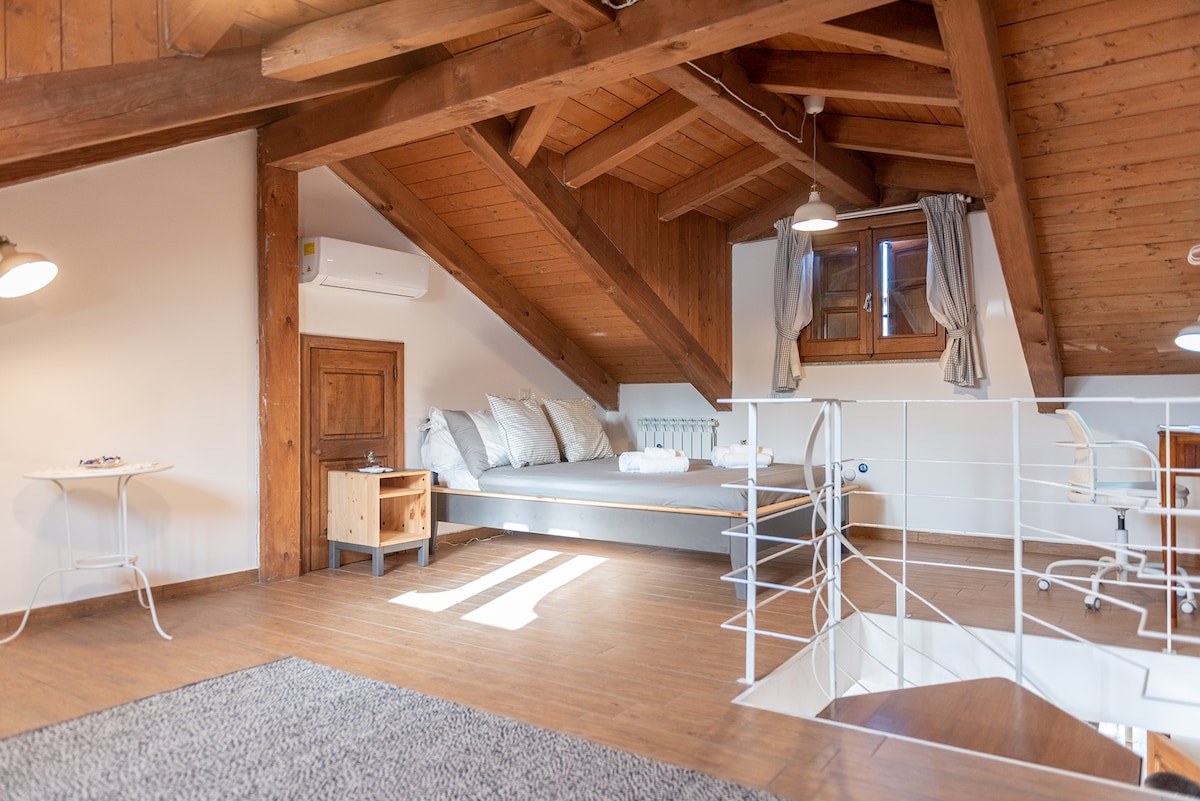 The bedroom features a comfortable bed with soft white linens, positioned under a wooden ceiling with exposed beams. Natural light filters through a window, creating a bright atmosphere. A small wooden nightstand and a workspace with a chair add functionality to the cozy space.
