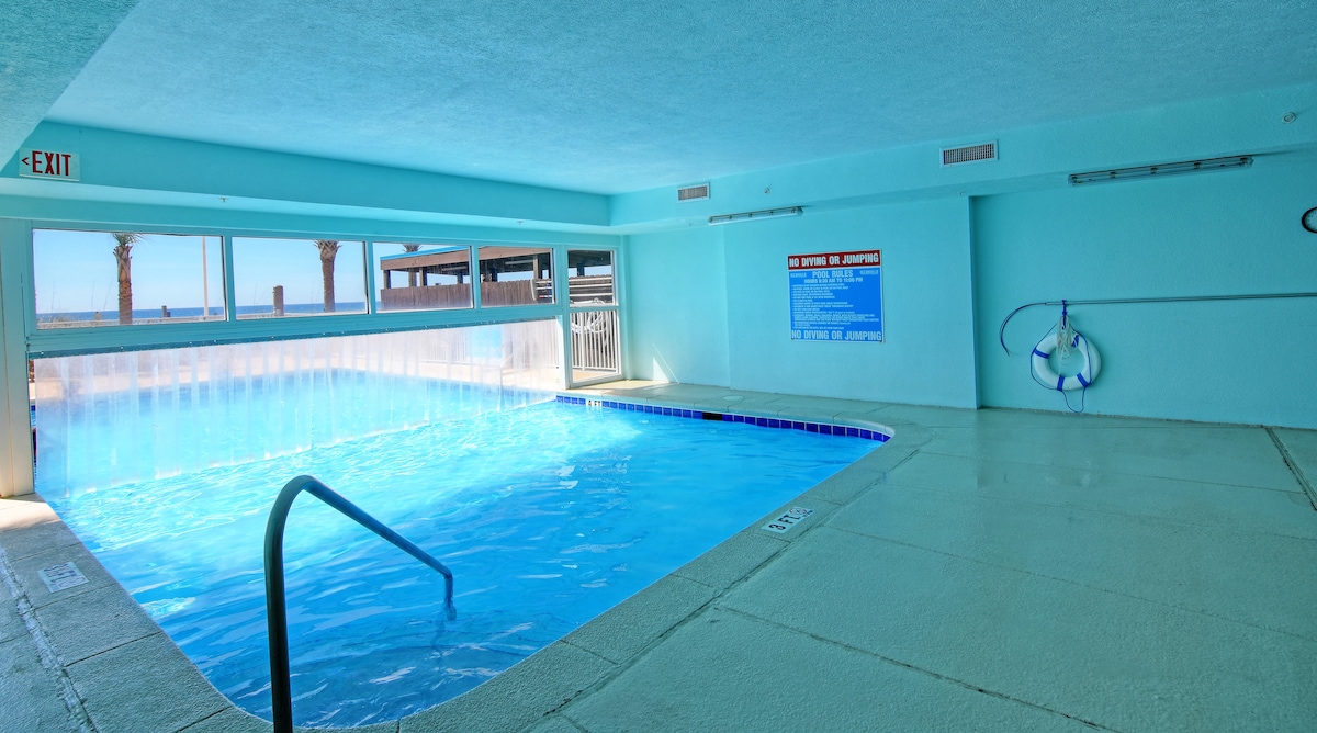 The indoor swimming pool area features a bright turquoise ambiance, with large windows offering views of the outdoor scenery. A gentle staircase provides access to the pool, which is lined with a clear blue water edge, enhancing the space’s inviting atmosphere.