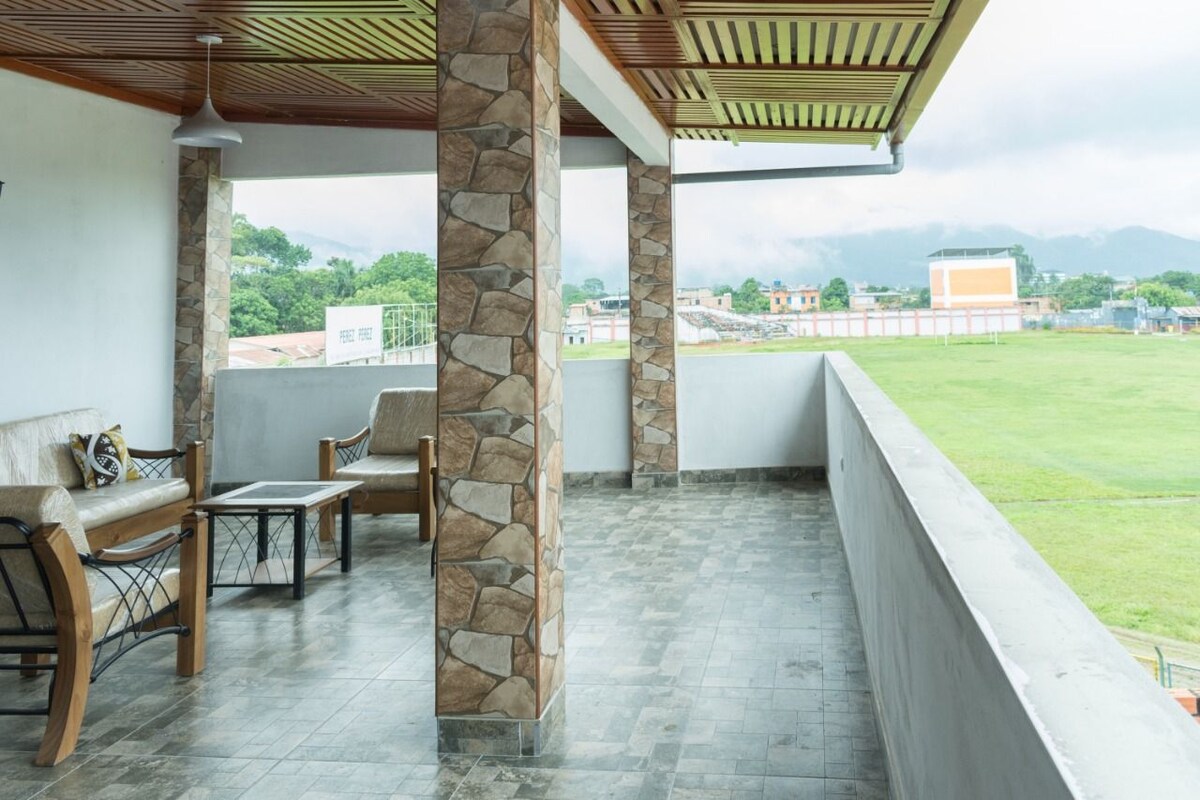 An extensive private terrace is shown, featuring stone pillars and a tiled floor. Comfortable seating arrangements include a woven sofa and wooden chairs, providing ample space for relaxation. A view of the surrounding greenery and distant mountains is available, contributing to a serene outdoor experience.