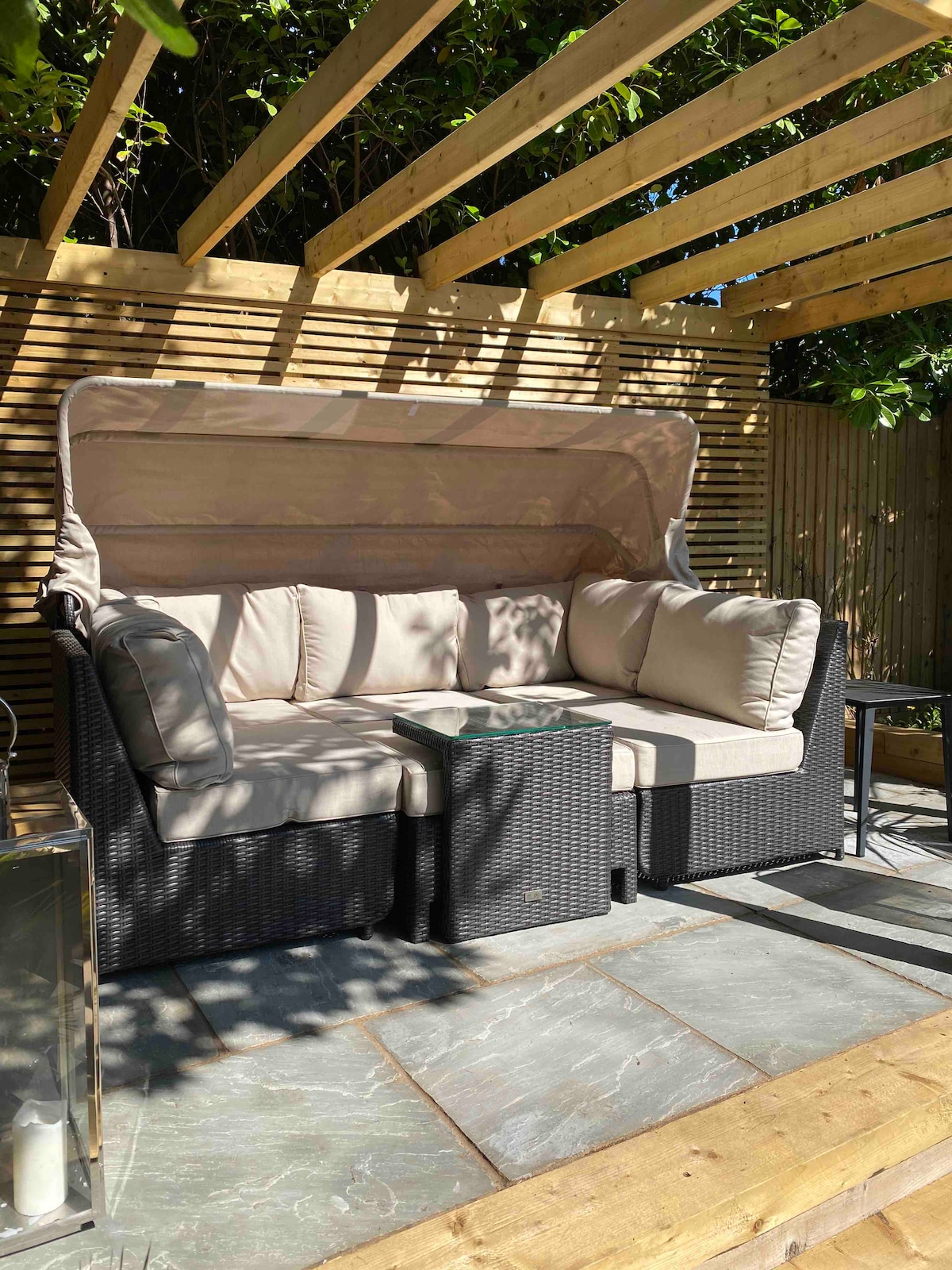 A comfortable seating area is featured with a sheltered sofa, upholstered in light fabric. The woven black rattan furniture is complemented by a small table, positioned on a stone floor surrounded by wooden slats and greenery.