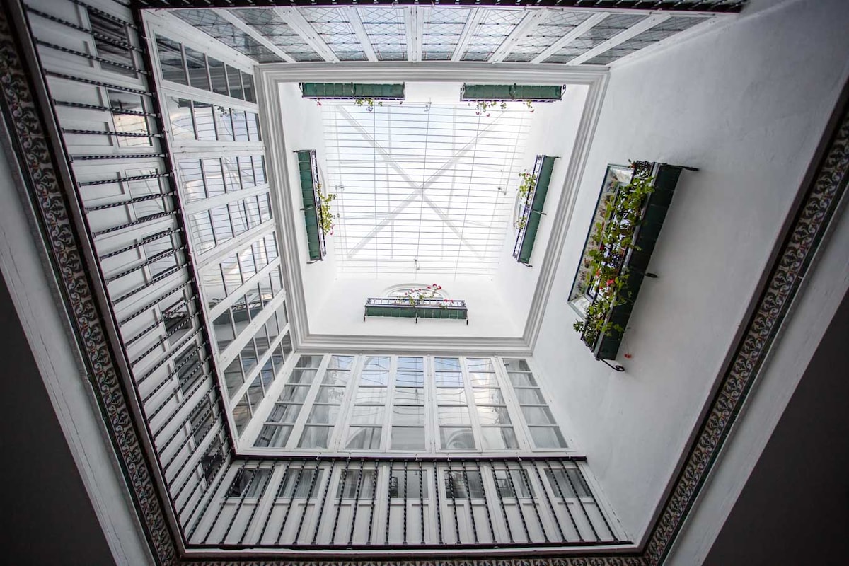 An atrium is highlighted by a glass skylight, allowing natural light to fill the space. Decorative metalwork frames large, symmetrical windows with potted plants visible at the edges. The walls are adorned with intricate tiles, creating a harmonious blend of light and design.