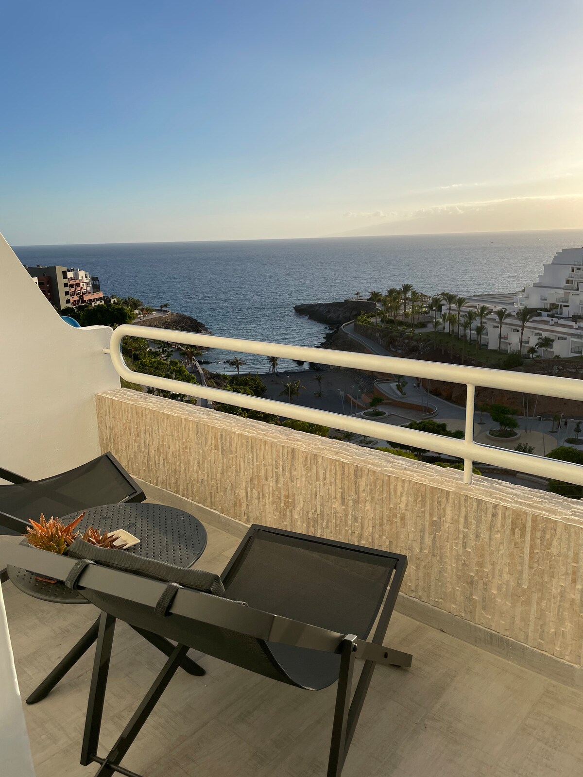 A small balcony features two black chairs oriented towards a stunning sea view. The coastline can be seen in the distance, with palm trees lining the area. Soft sunlight casts a warm glow, enhancing the overall tranquil atmosphere.