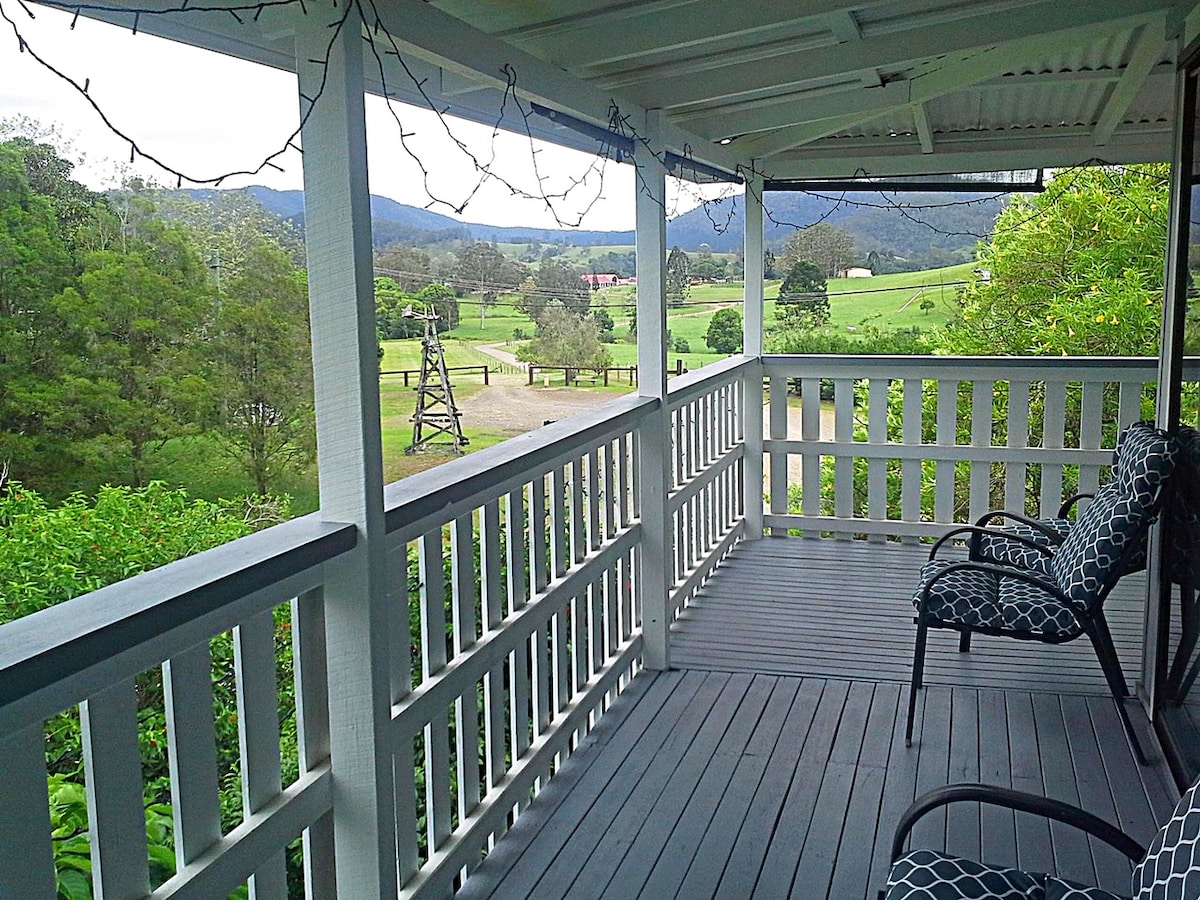 A covered porch is presented with decorative string lights overhead. Two patterned chairs offer a comfortable spot to enjoy views of rolling green hills and distant mountains. Open spaces and a rustic structure can be seen in the landscape beyond.