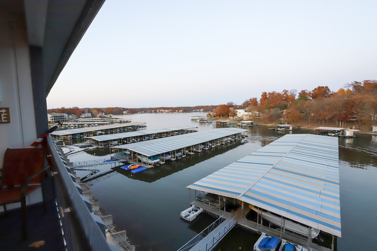 A calm waterway is observed from the balcony, featuring several boat docks with covered slips. Autumn foliage lines the shore, showcasing trees in warm hues. The scene captures the serenity of the water, reflecting the tranquil ambiance of the surrounding area.
