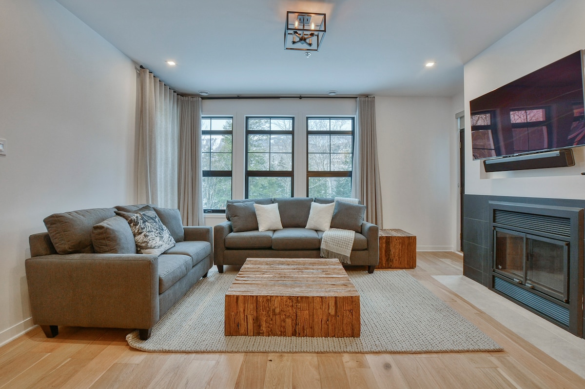 A modern living area features two gray sofas positioned around a wooden coffee table on a light area rug. Large windows allow natural light to fill the space, with sheer curtains softly framing the views. A contemporary fireplace is mounted on the wall opposite the seating.