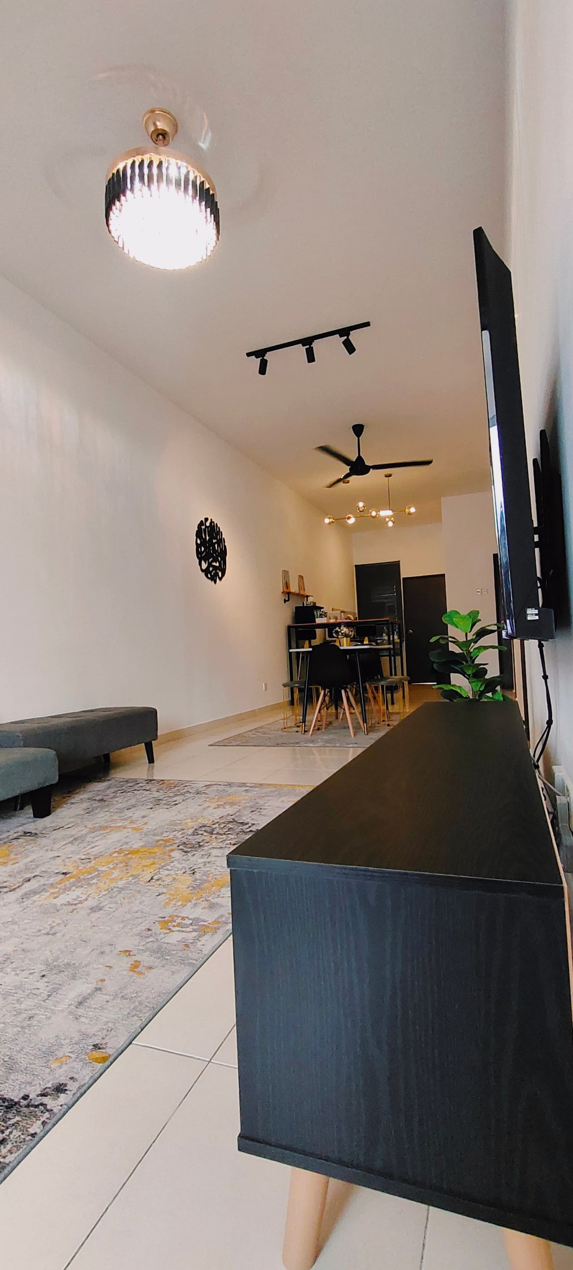 The living area features a low-profile black TV cabinet and a plush gray sofa, positioned along a sleek, light-toned wall. A dining table and chairs are visible in the background, and soft lighting is provided by a stylish ceiling lamp and track lights.