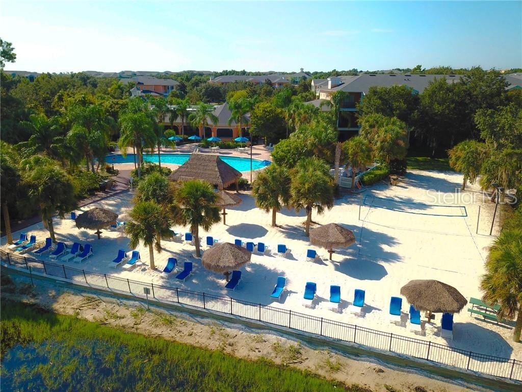 An aerial view captures the sandy beach area lined with palm trees and shaded cabanas. Lounge chairs are arranged neatly in front of the beach, while a pool area with vibrant blue water is visible in the background surrounded by lush landscaping.