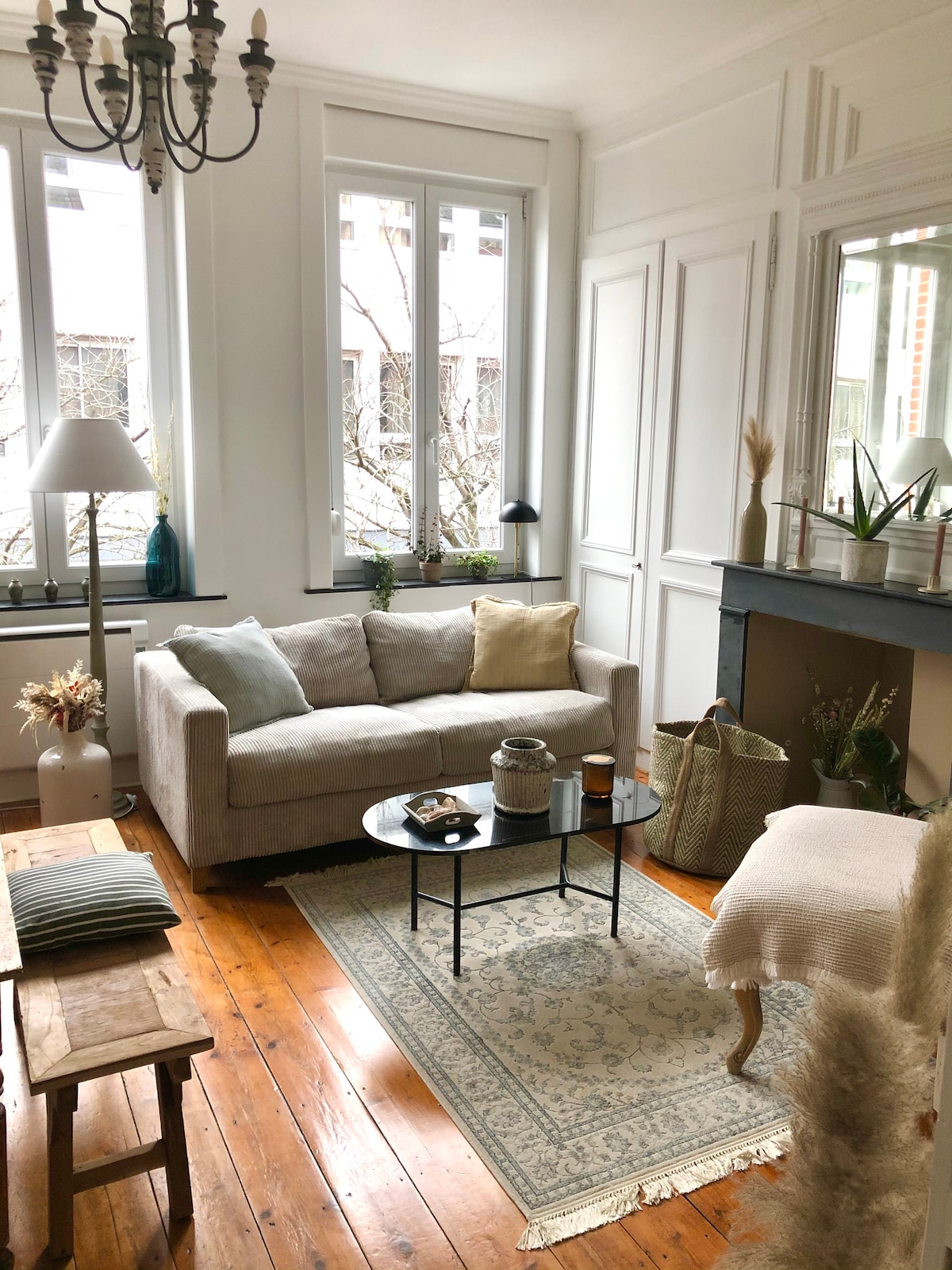 A light-filled living area features a soft, neutral-colored couch paired with a low coffee table. Decor includes a woven basket, decorative pillows, and potted plants. Large windows allow natural light to illuminate the wooden flooring and subtle area rug.
