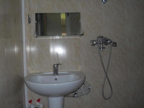 A clean bathroom space features a white sink positioned beneath a wall-mounted mirror. A showerhead is mounted on the wall, with a flexible hose attached for convenience. Light-colored tile surrounds the area, contributing to a bright and functional environment.