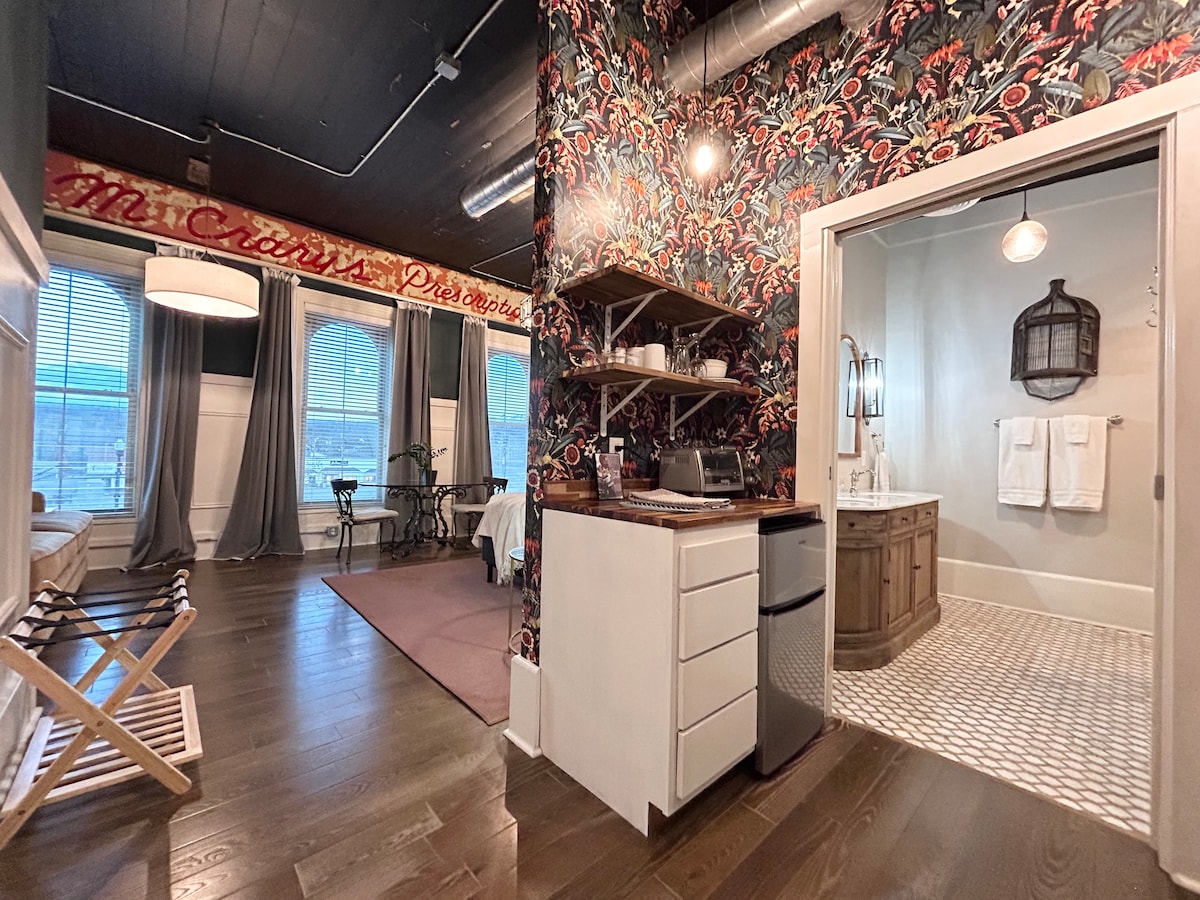 The living area showcases a kitchenette with a refrigerator and microwave, paired with open shelving. A large bathroom leads off the space, featuring double vanities and a walk-in shower. Light streams through tall windows, creating an inviting ambiance throughout the suite.