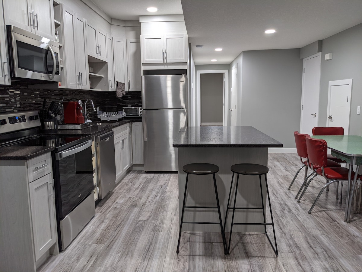 A modern kitchen showcases stainless steel appliances, including an oven and refrigerator, complemented by sleek cabinetry and a stylish backsplash. A central island with two bar stools provides additional seating. The dining area features a glass table surrounded by red chairs, enhancing the space's contemporary design.