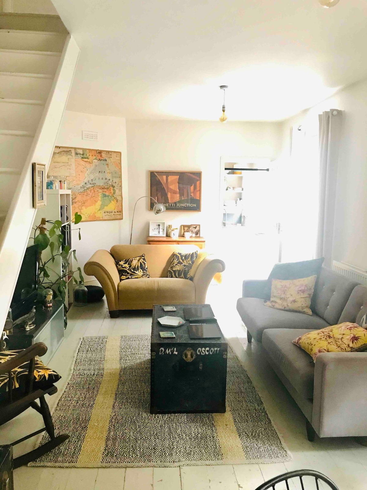 A comfortable living area is featured, showcasing two sofas in neutral tones. A vintage-style coffee table sits at the center, surrounded by potted plants. Natural light enters through large windows, illuminating the space and highlighting framed artworks on the walls.