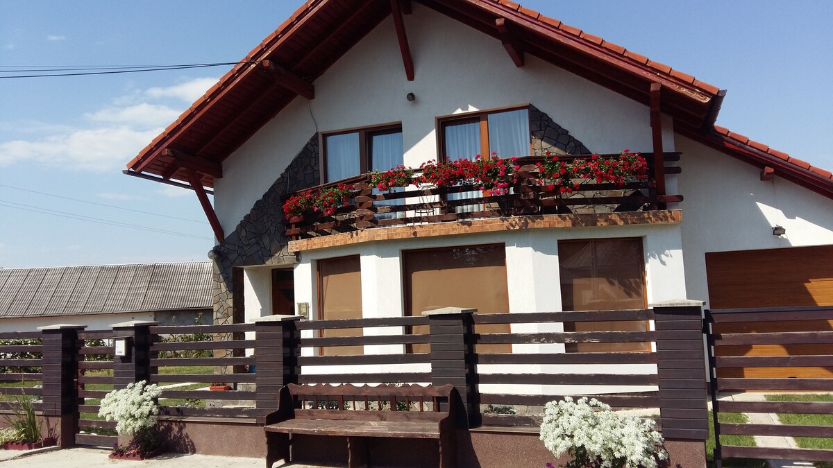 The exterior of a two-story house features a combination of stone and wooden elements. Flower boxes with vibrant blooms adorn the balcony. A wooden bench sits in front of the home, complemented by neatly arranged plants along the pathway. The sky is clear and sunny.