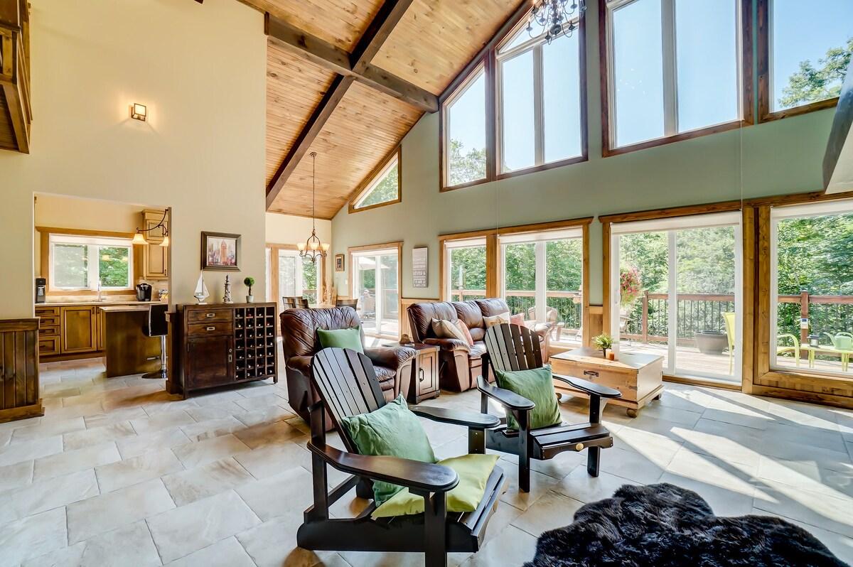 A spacious living area features large windows allowing natural light to fill the room. Wooden beams enhance the height of the ceiling, while comfortable seating arrangements are complemented by green accent pillows. A well-appointed kitchen is visible in the background, leading to a large outdoor patio.